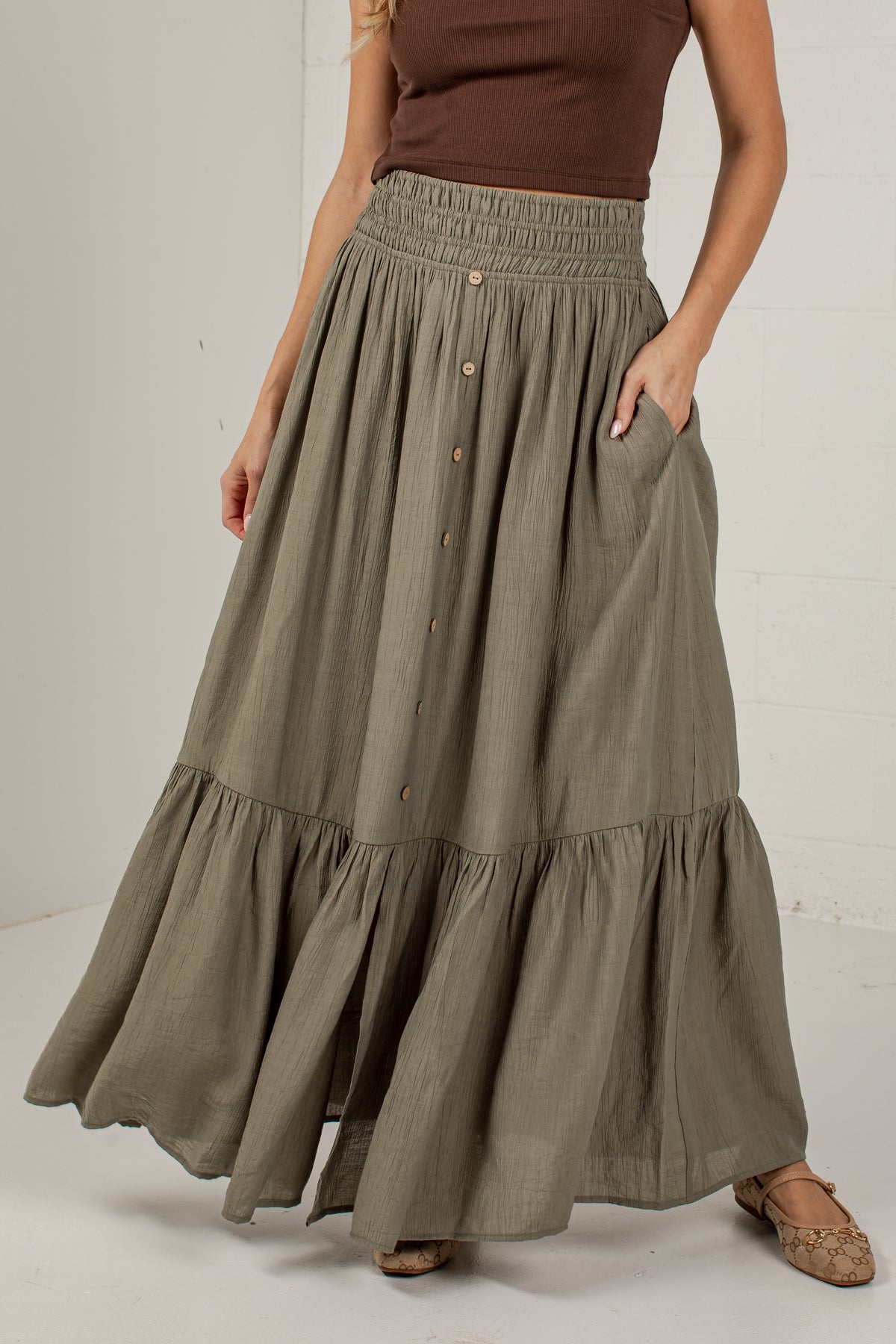 A woman wearing a long, tiered, high-waisted maxi skirt in a muted olive green color. The skirt features a button-down front, side pockets, and a gathered elastic waistband.