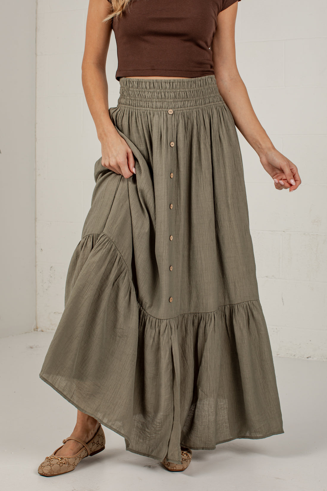 A woman wearing a long, tiered, button-front maxi skirt in a muted sage green color, paired with a brown sleeveless top and brown flat shoes, standing against a white wall.