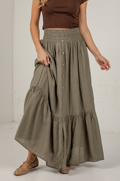 A woman wearing a long, tiered, button-front maxi skirt in a muted sage green color, paired with a brown sleeveless top and brown flat shoes, standing against a white wall.