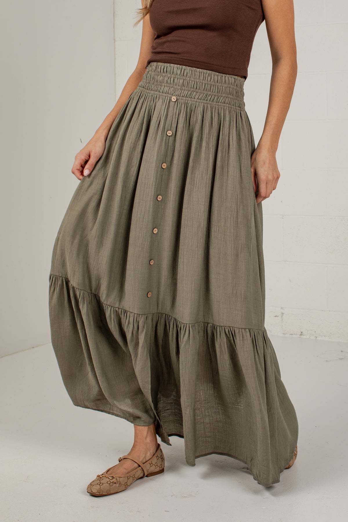 A woman wearing a high-waisted, tiered maxi skirt in a muted olive green color. The skirt features a smocked waistband, a vertical row of brown buttons down the front, and a flared, voluminous silhouette.