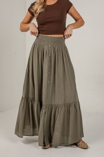 A woman wearing a brown short-sleeve crop top and a high-waisted, tiered olive green maxi skirt with front buttons, standing against a white studio wall.