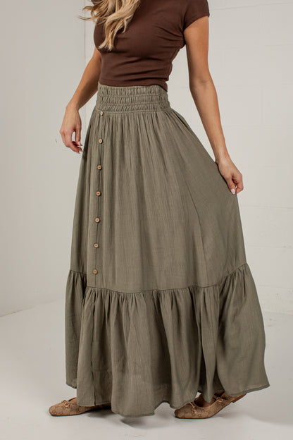 A woman wearing a high-waisted, tiered maxi skirt in a muted olive green color, paired with a brown top and brown shoes, shown from the waist down against a light gray studio background.