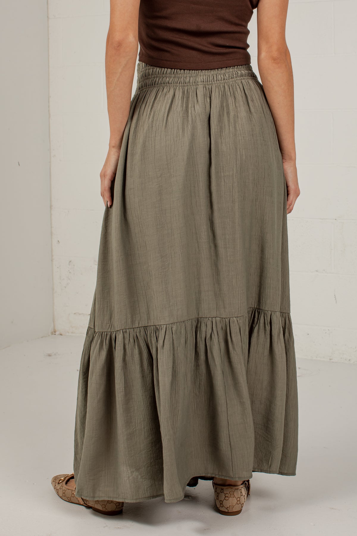 A woman wearing a long, tiered maxi skirt in a muted sage green color, paired with a brown top and cork sandals, standing against a white studio wall.