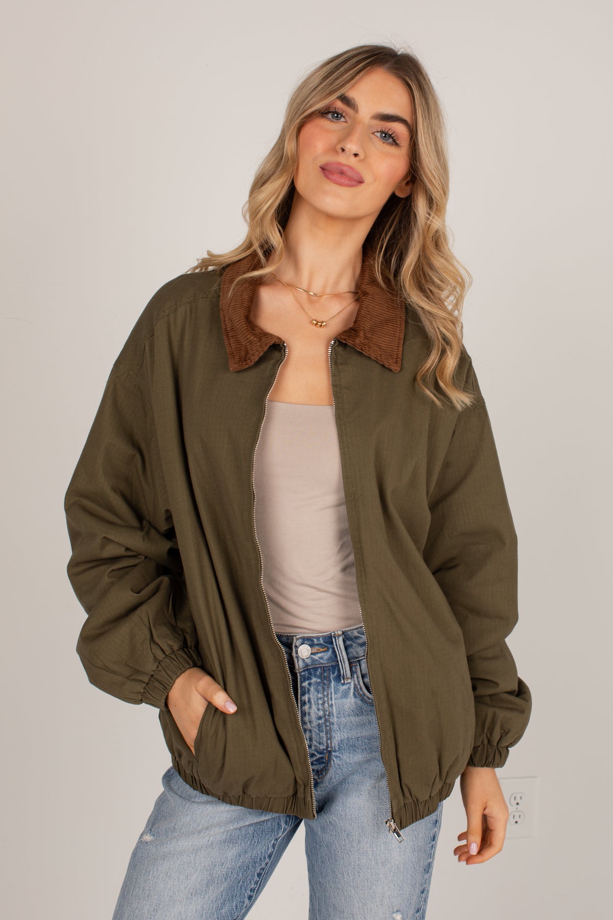A woman wearing an olive green zip-up jacket with a brown suede-like collar, styled over a beige tank top and blue jeans, photographed against a neutral light gray background.