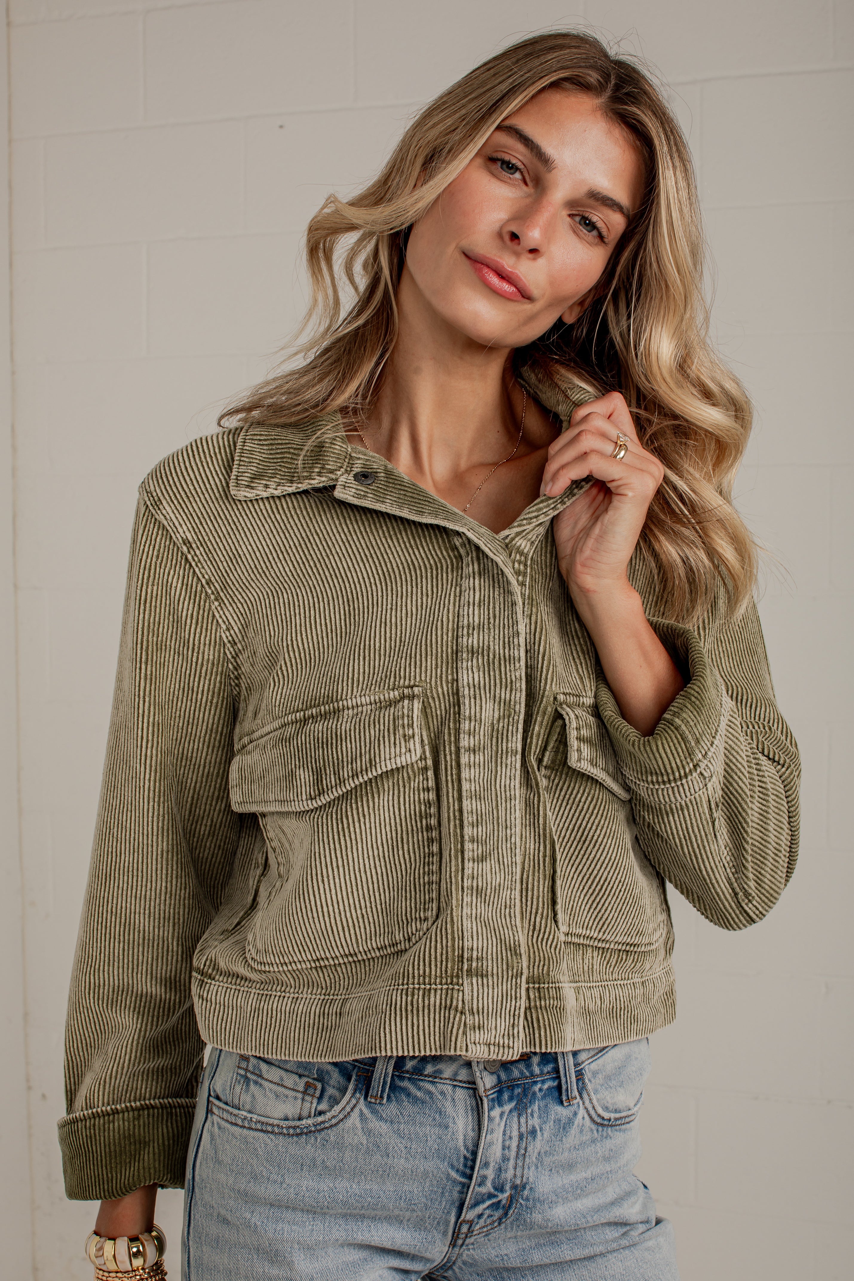 A woman wearing a cropped corduroy jacket in a muted sage green color, paired with light-wash blue jeans, posing against a light gray brick wall.