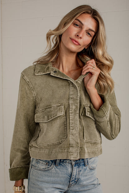 A woman wearing a cropped corduroy jacket in a muted sage green color, paired with light-wash blue jeans, posing against a light gray brick wall.