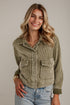 A woman wearing a cropped corduroy jacket in a muted sage green color, paired with light-wash blue jeans, standing against a light-colored brick wall.