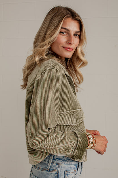 A woman wearing a sage green corduroy jacket and light blue jeans, photographed from the side against a light gray brick wall.