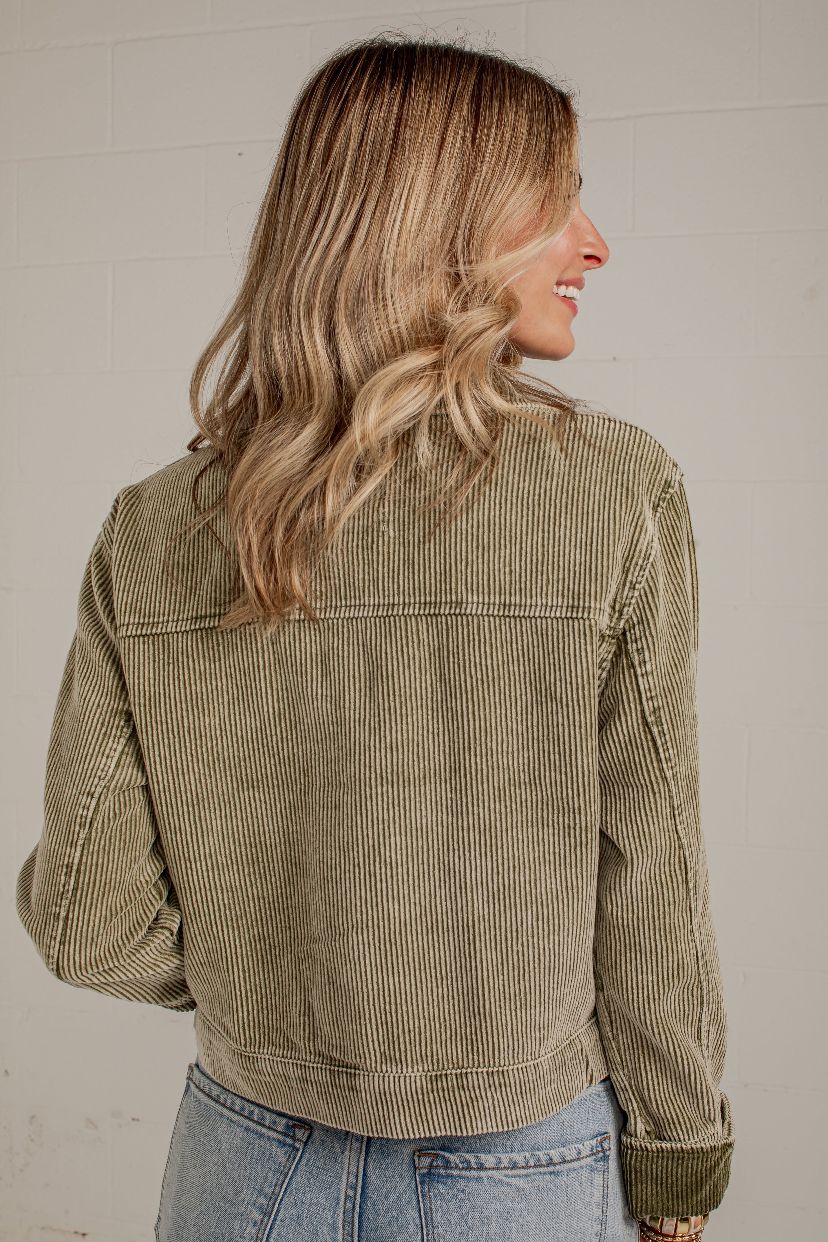 A woman shown from the back wearing an olive green corduroy jacket and light blue jeans, standing against a white brick wall.