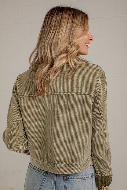 A woman shown from the back wearing an olive green corduroy jacket and light blue jeans, standing against a white brick wall.