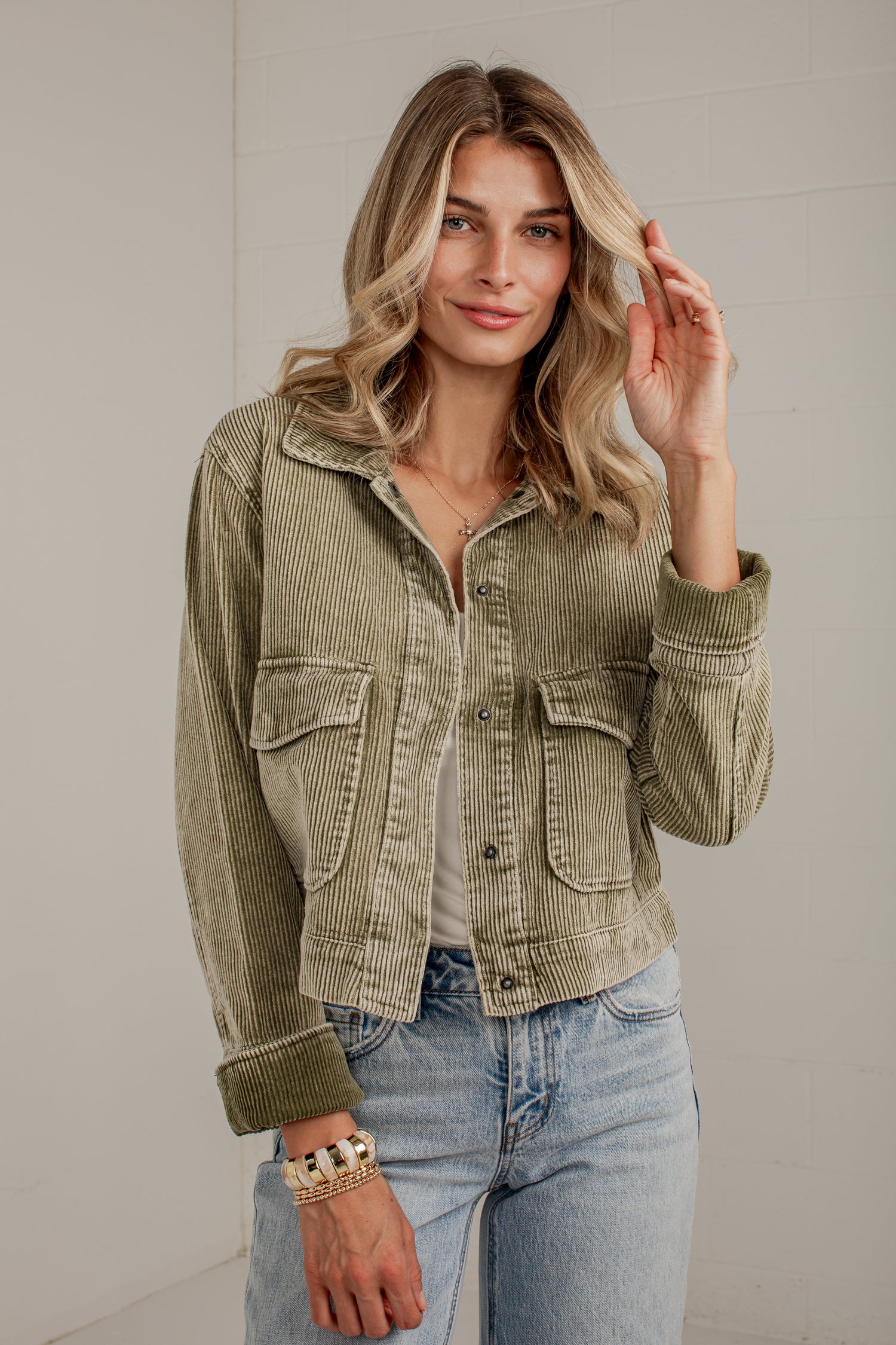 A woman wearing a cropped olive green corduroy jacket with front flap pockets, styled with light blue jeans and a gold bracelet, standing against a light gray wall.