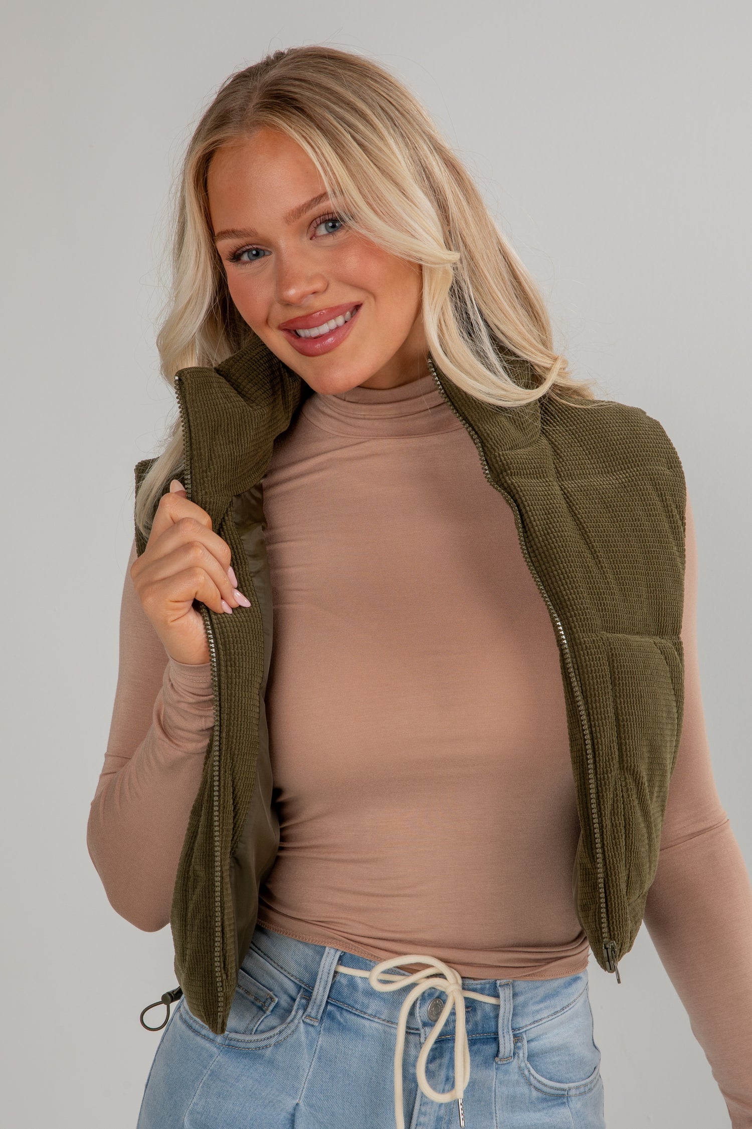 Woman wearing a green vest over a brown top and blue jeans on a plain background