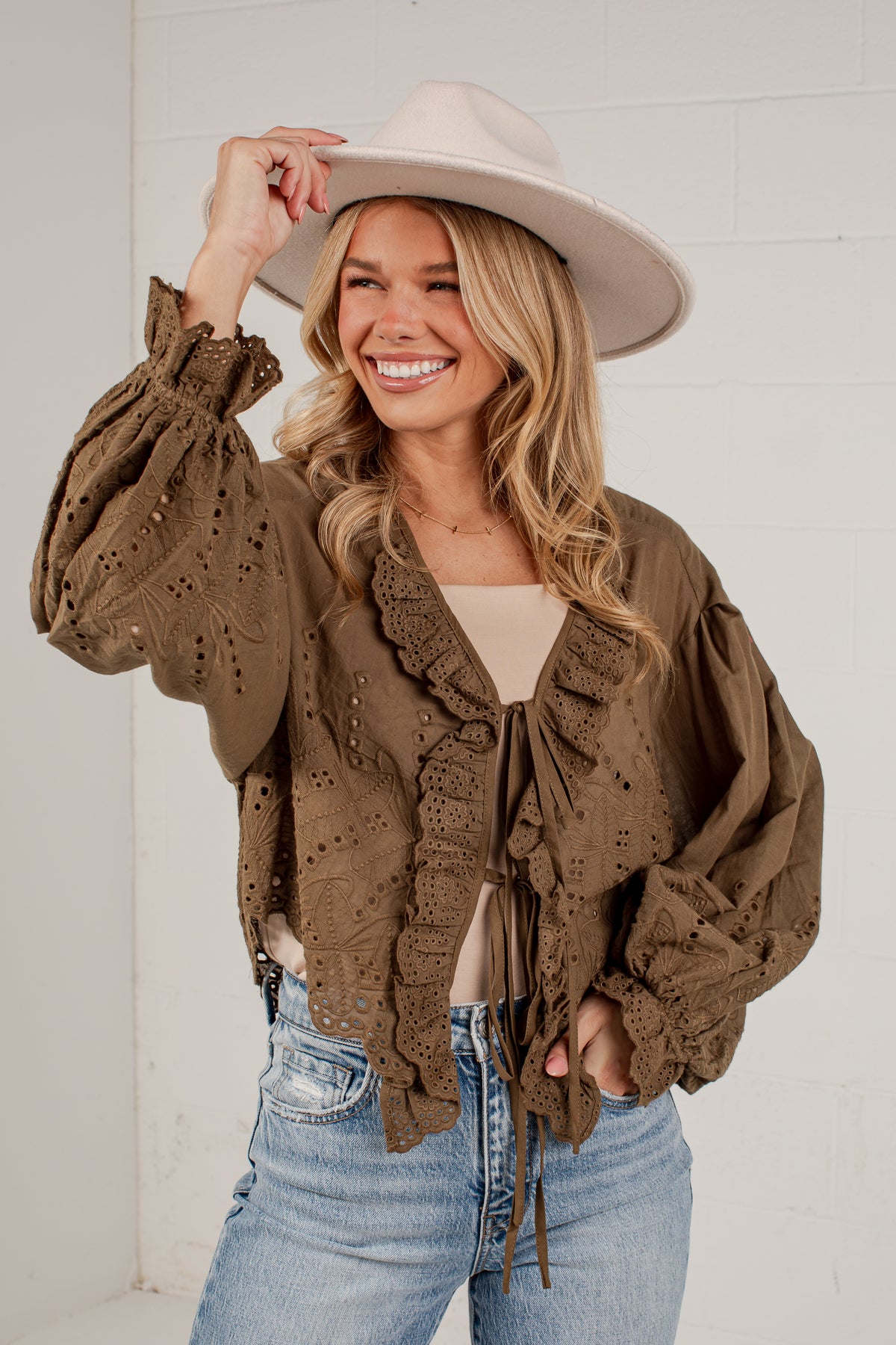 A woman wearing a brown eyelet lace cropped jacket with ruffle trim, a beige top, blue jeans, and a wide-brimmed beige hat, posing against a white brick wall.