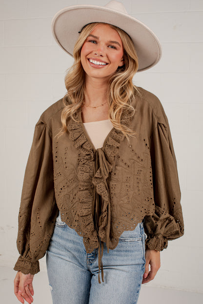 A woman wearing a brown eyelet lace blouse with ruffles and long puff sleeves, paired with a wide-brim beige hat and light blue jeans, standing against a white brick wall.