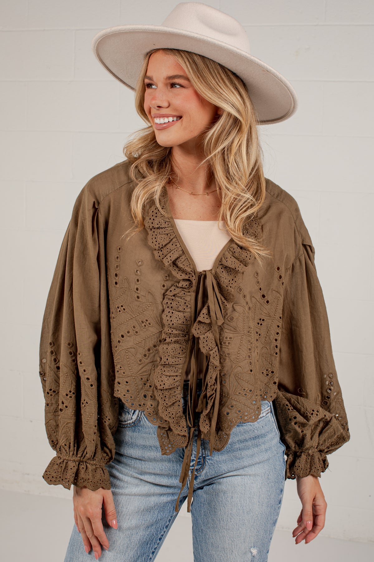 A woman wearing a brown eyelet blouse with ruffles and tie-front detailing, paired with light blue jeans and a wide-brim beige hat, standing against a white brick wall.