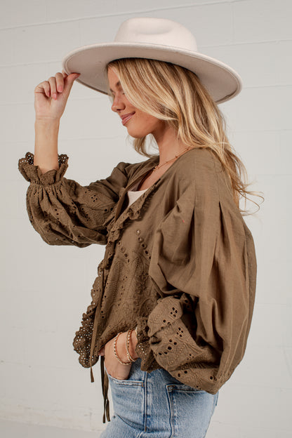 A woman wearing a brown eyelet blouse with ruffled trim, blue jeans, and a wide-brimmed cream hat, posing against a white paneled wall.