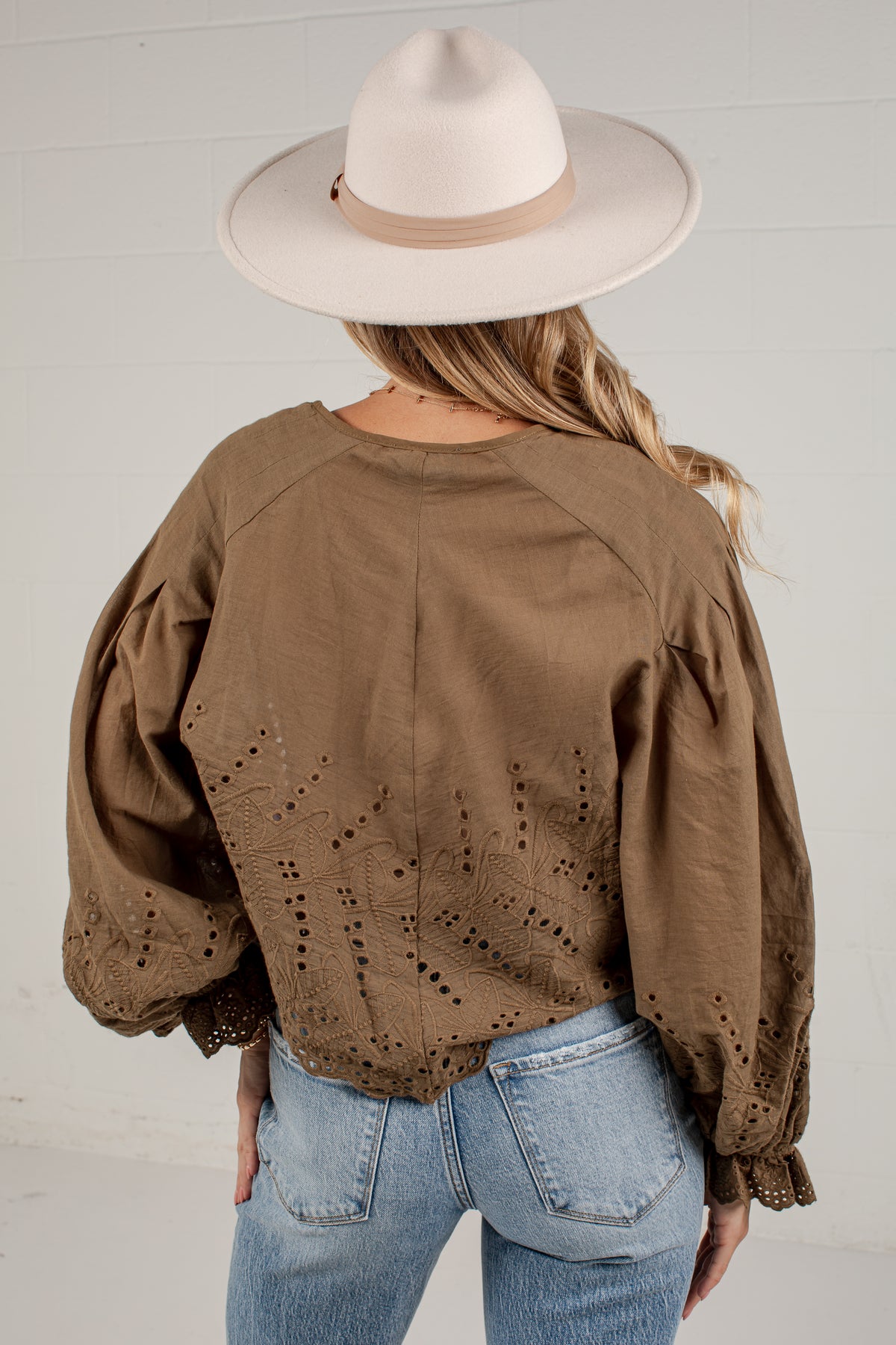 A woman viewed from behind wearing a brown eyelet blouse with voluminous sleeves, paired with light blue jeans and a cream wide-brim hat against a white paneled wall.