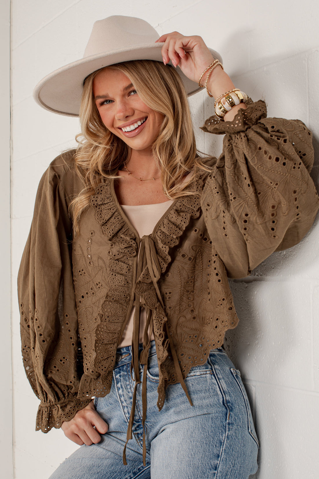 A woman wearing a brown eyelet lace long-sleeve blouse with ruffles and tie details, paired with a beige wide-brim hat and light blue jeans, posing against a white brick wall.