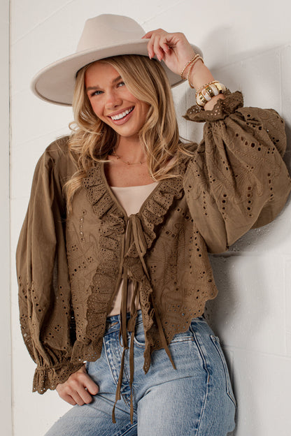 A woman wearing a brown eyelet lace long-sleeve blouse with ruffles and tie details, paired with a beige wide-brim hat and light blue jeans, posing against a white brick wall.