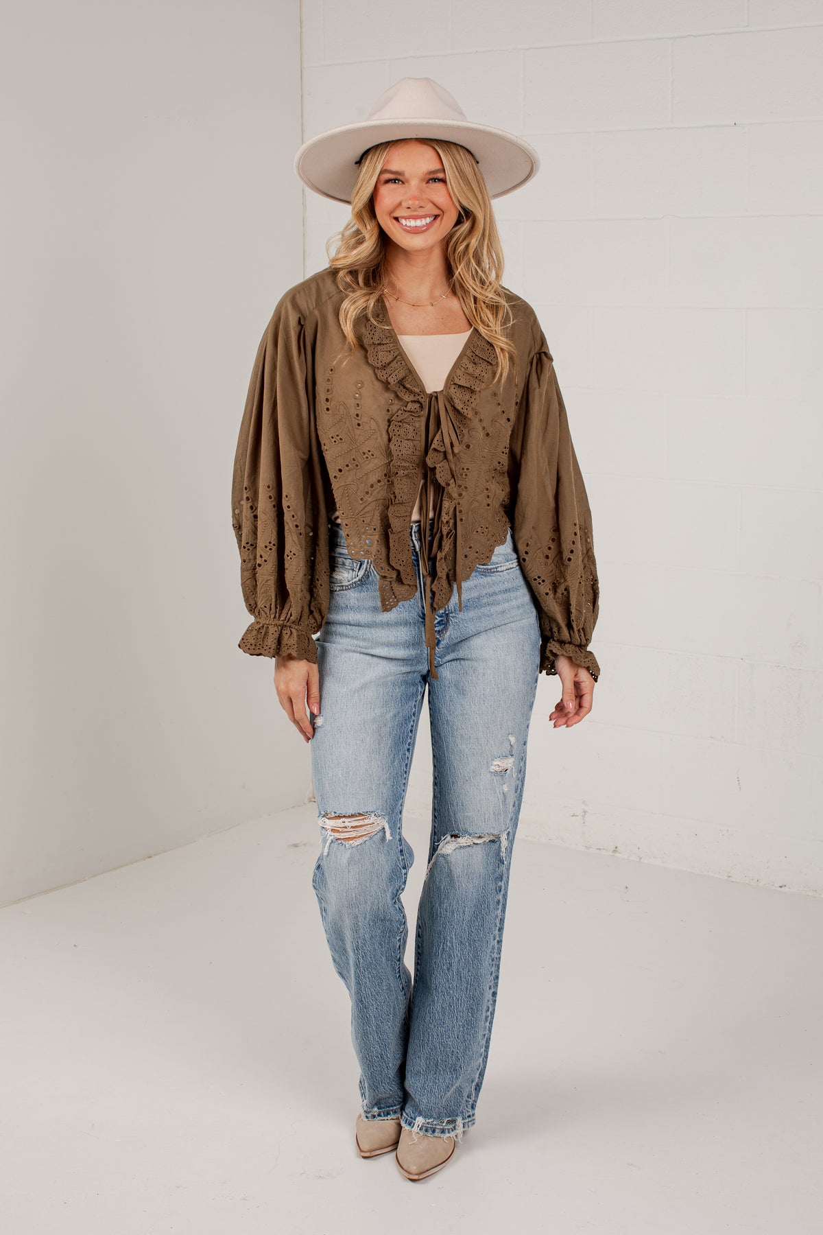 A woman modeling a brown ruffled long-sleeve blouse with a tie waist, paired with light-wash distressed jeans, beige heels, and a wide-brim beige hat, standing against a white studio wall.