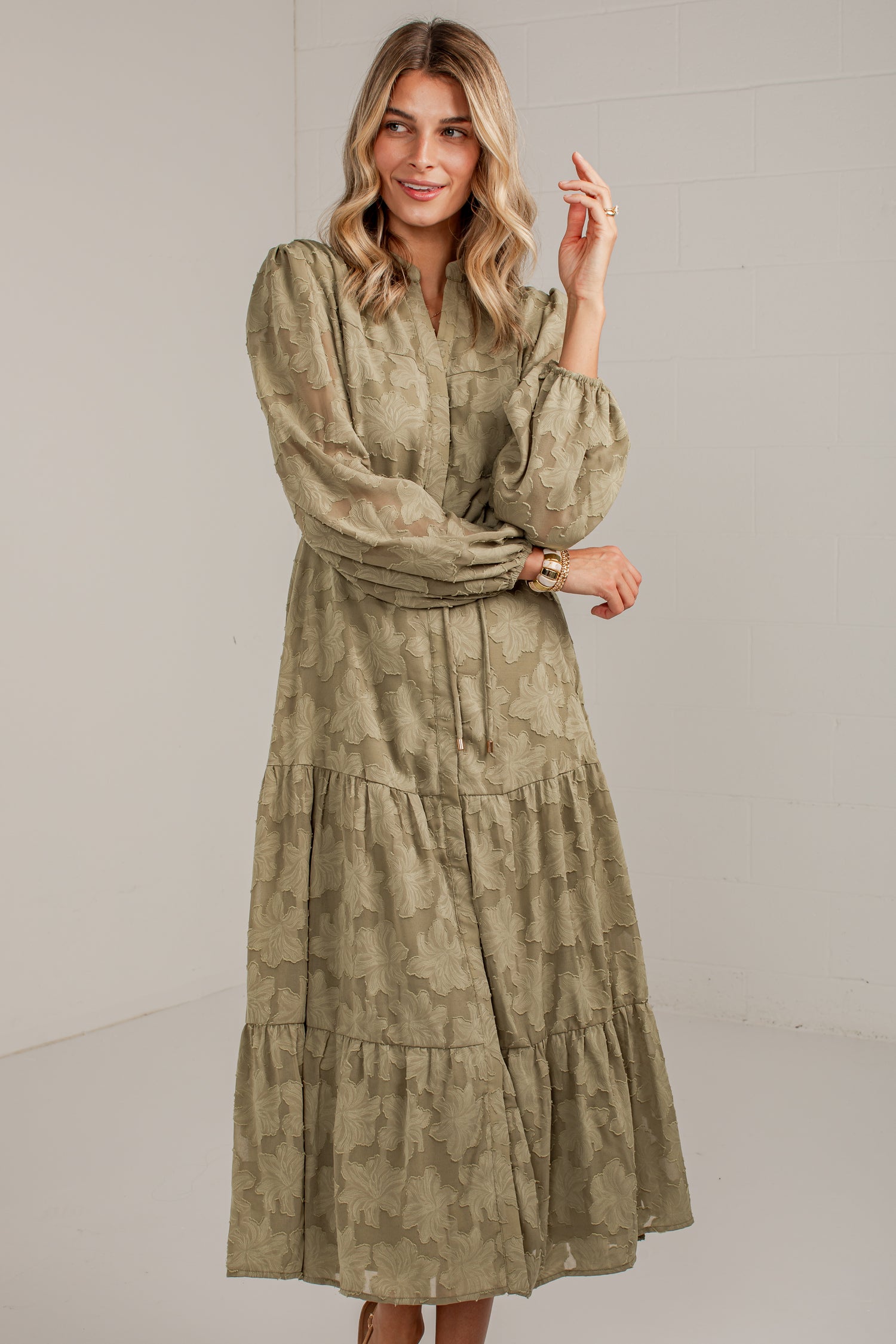 A woman modeling a long-sleeved, tiered midi dress with a muted olive green and beige abstract pattern, set against a plain light gray studio background.