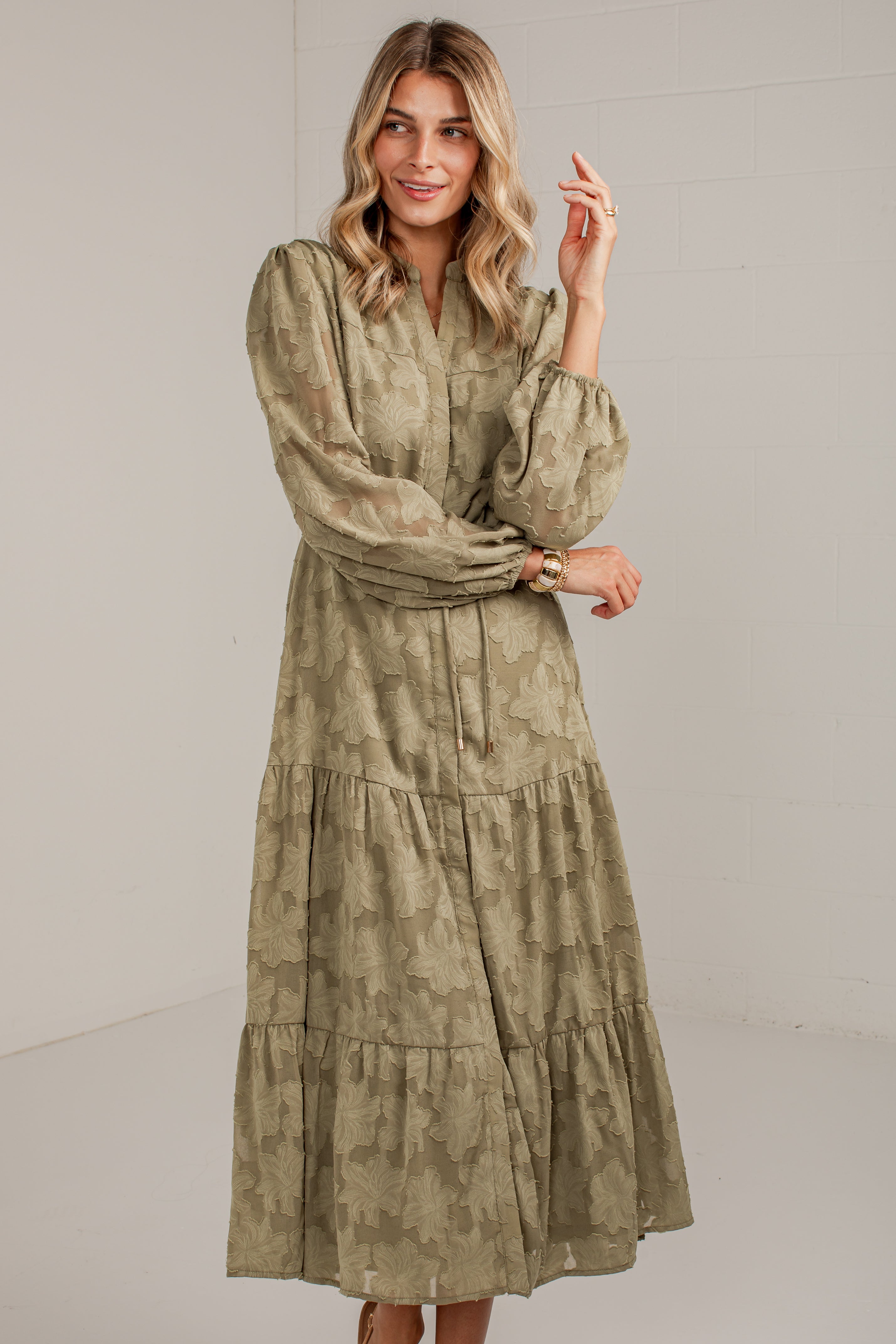A woman modeling a long-sleeved, tiered midi dress with a muted olive green and beige abstract pattern, set against a plain light gray studio background.