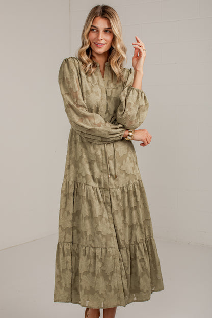 A woman modeling a long-sleeved, tiered midi dress with a muted olive green and beige abstract pattern, set against a plain light gray studio background.