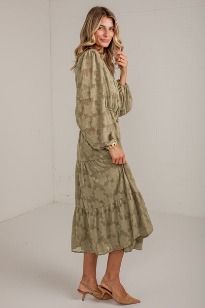 A woman modeling a long-sleeve midi dress with a muted olive green and beige abstract print, paired with tan slingback heels, in a bright studio setting.