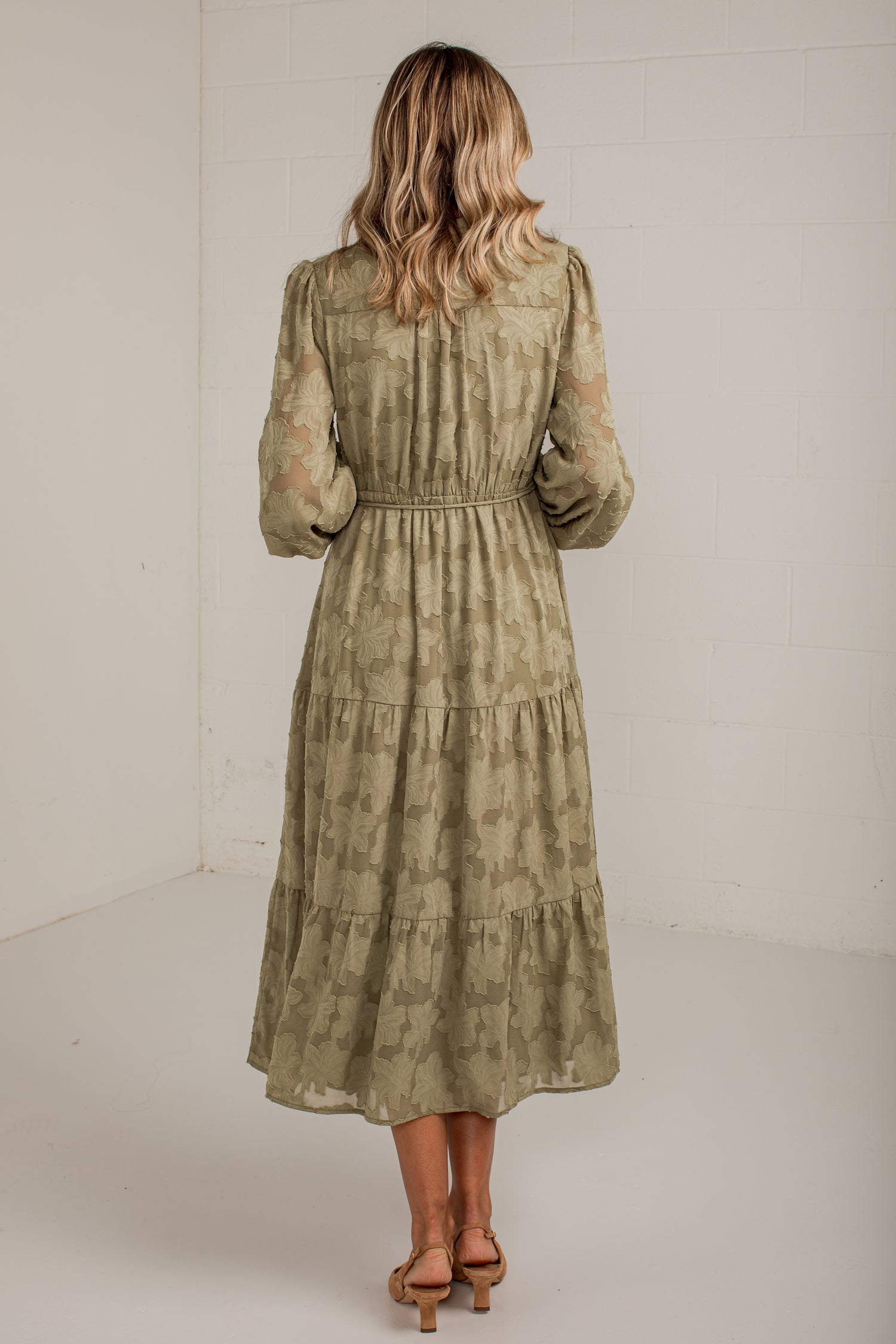 A woman viewed from behind wearing a long-sleeve, tiered midi dress in a muted olive green and beige patterned fabric, paired with tan heeled sandals, set against a plain white studio background.