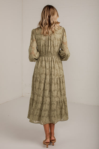 A woman viewed from behind wearing a long-sleeve, tiered midi dress in a muted olive green and beige patterned fabric, paired with tan heeled sandals, set against a plain white studio background.