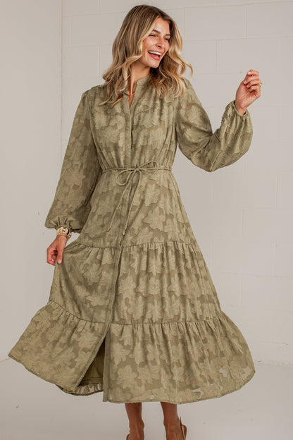 A woman modeling a long-sleeved, tiered midi dress in a muted olive green and beige tie-dye pattern, paired with tan pointed-toe heels, against a white studio background.