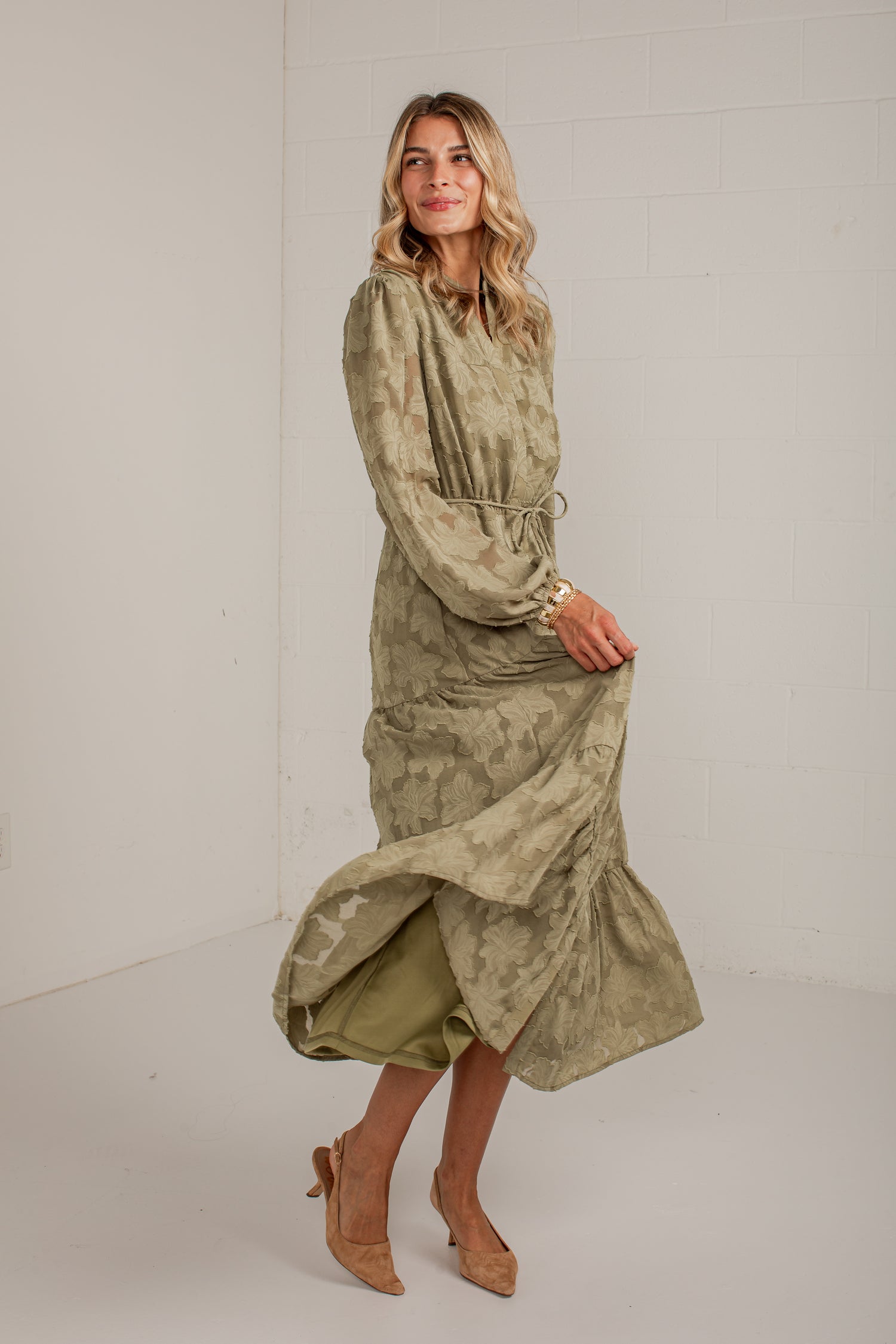 A woman modeling a long-sleeved, high-neck midi dress with a textured, mottled olive green and beige pattern. The dress features a drawstring waist and is styled with tan pointed-toe heels in a bright, neutral studio setting.