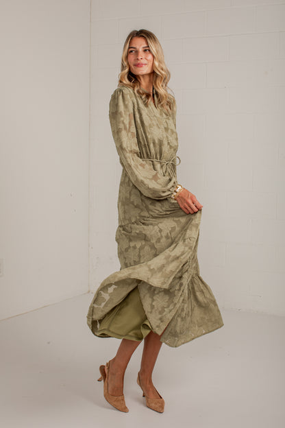 A woman modeling a long-sleeved, high-neck midi dress with a textured, mottled olive green and beige pattern. The dress features a drawstring waist and is styled with tan pointed-toe heels in a bright, neutral studio setting.