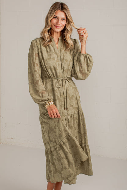 A woman modeling a long-sleeved olive green and beige patterned midi dress with a tie waist, standing in a bright white studio setting.