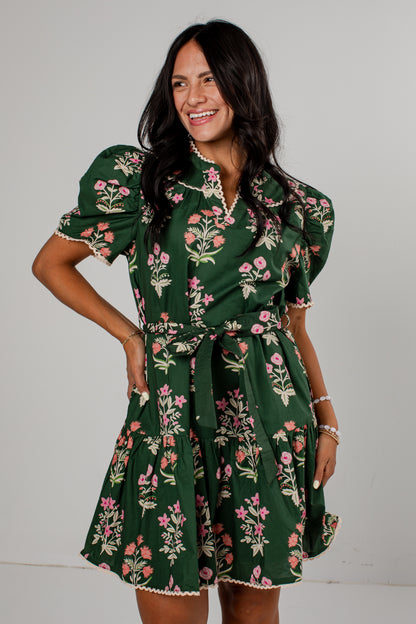 A woman modeling a dark green short-sleeve mini dress with a pink and white floral print, featuring puff sleeves, a collar, and a matching fabric tie belt, set against a plain light gray background.