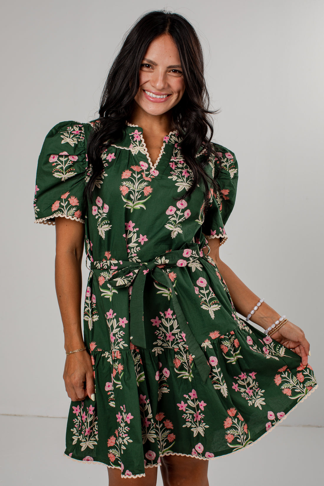 A woman modeling a dark green short-sleeve mini dress with a pink and beige floral print, featuring puff sleeves, a V-neckline, and a matching fabric tie belt.