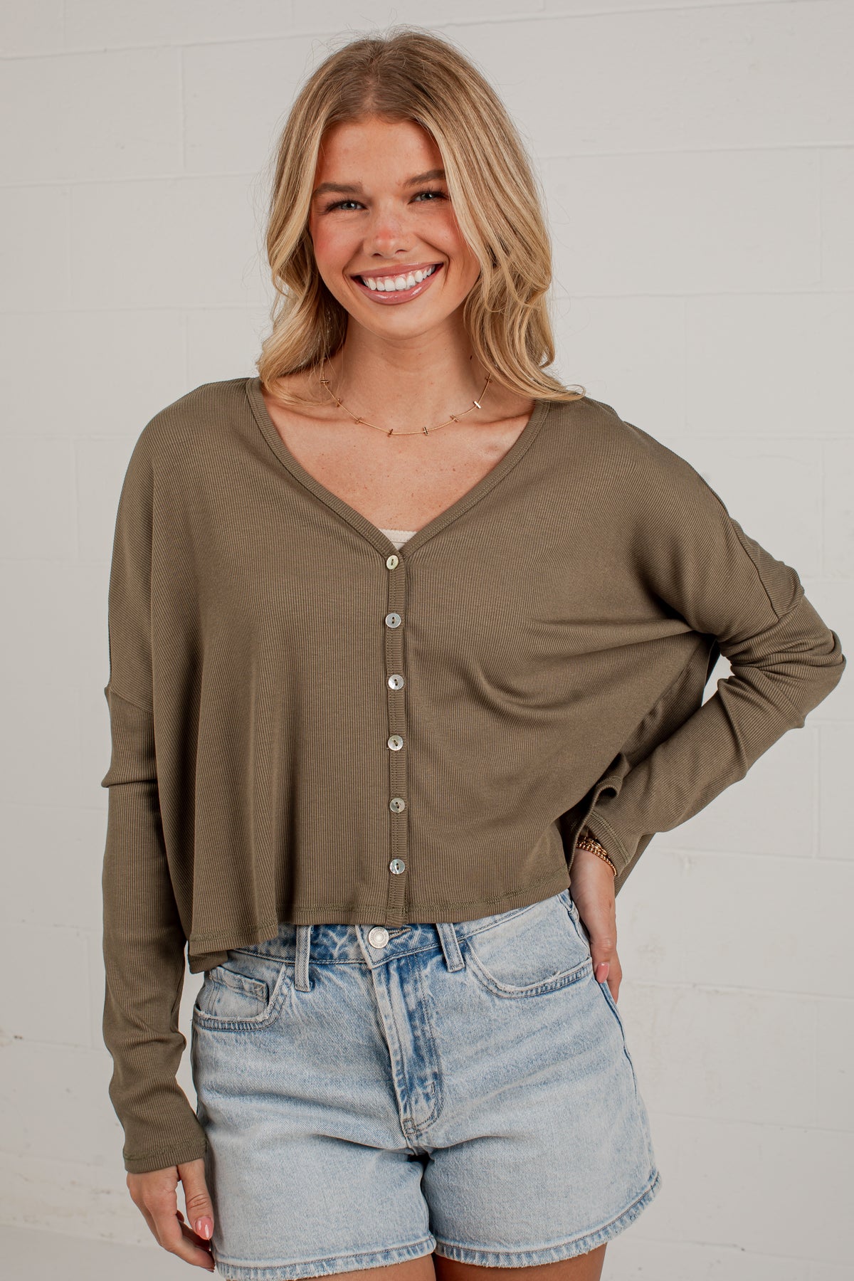 A smiling blonde woman wearing a loose, long-sleeve olive-brown button-front top and light-wash denim shorts, standing against a white brick wall.