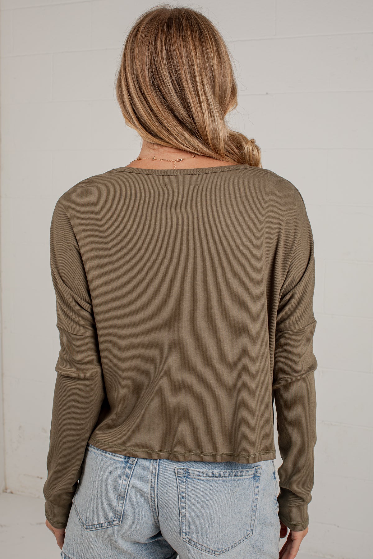 A woman viewed from behind wearing a cropped, long-sleeve ribbed knit top in a muted olive brown color, paired with light-wash blue jeans against a white brick wall.