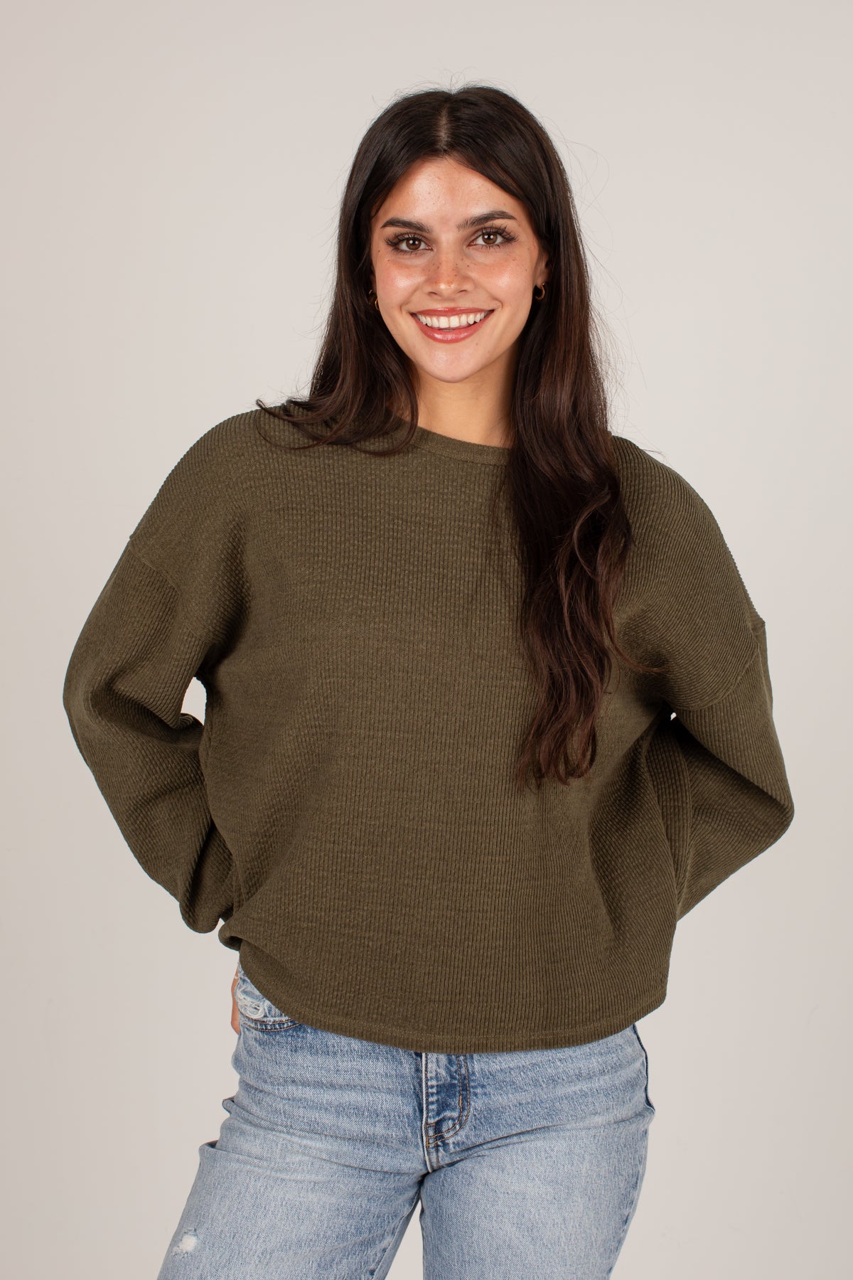 A woman with long dark hair wearing an olive green waffle-knit sweater and light blue jeans, standing against a plain light gray studio background.