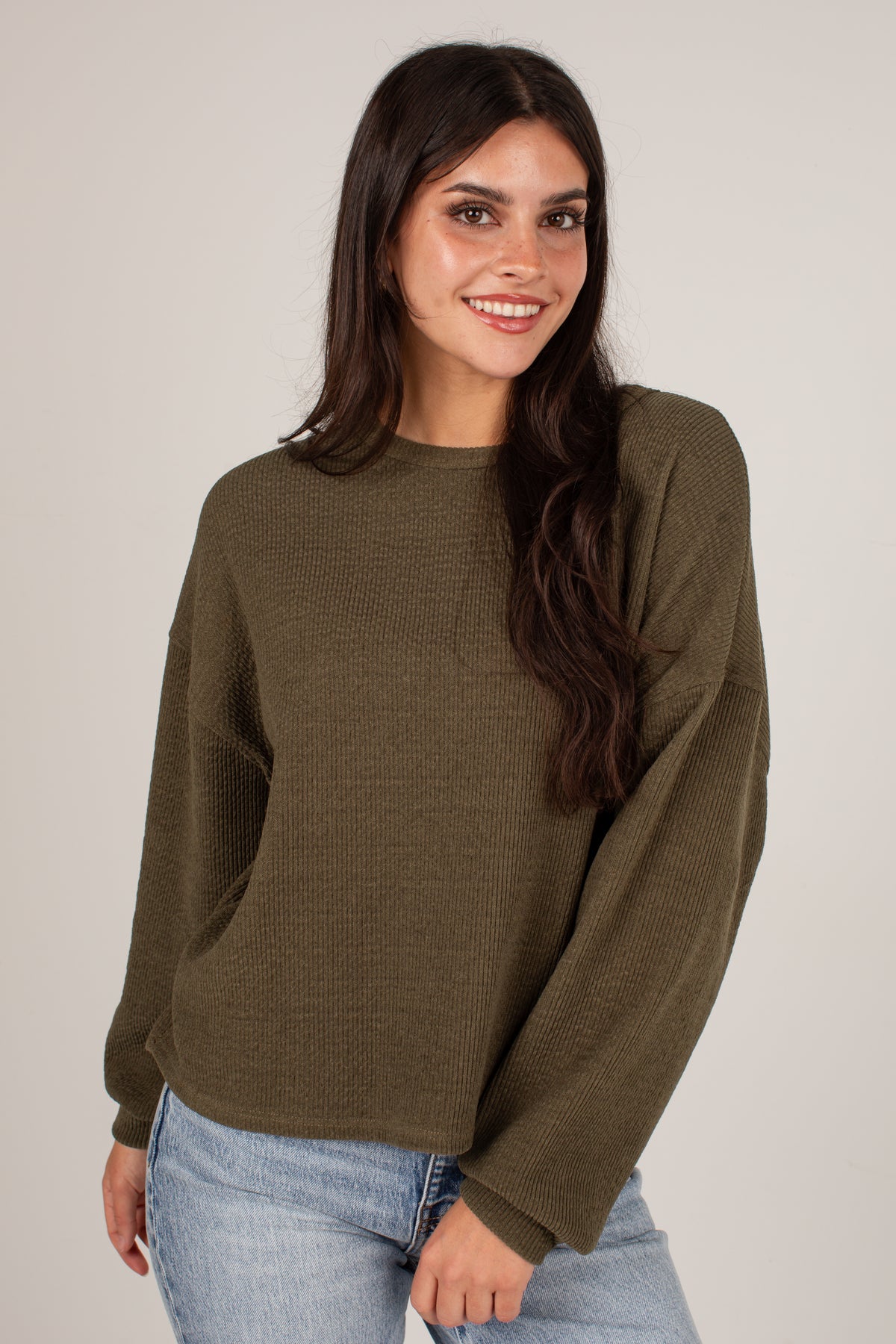 A woman with long dark hair wearing an olive green long-sleeve knit sweater and light blue jeans, standing against a plain light gray studio background.