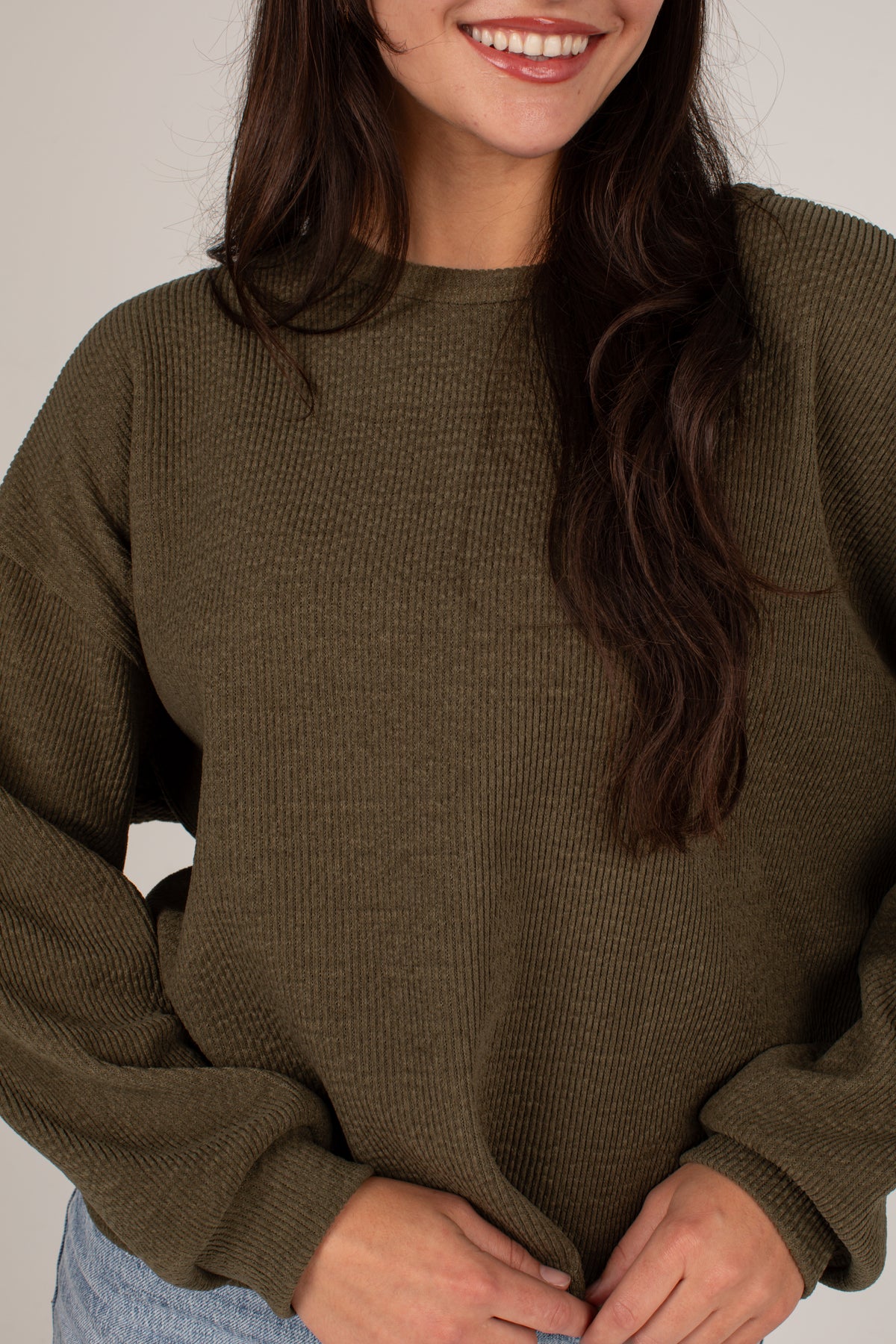 A woman wearing a ribbed knit crewneck sweater in olive green, shown from the chest down against a plain light background.