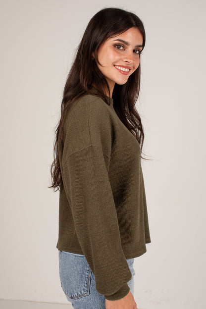 A woman with long dark hair is shown from the side/back wearing an olive green ribbed knit sweater and light blue jeans against a plain light gray studio background.
