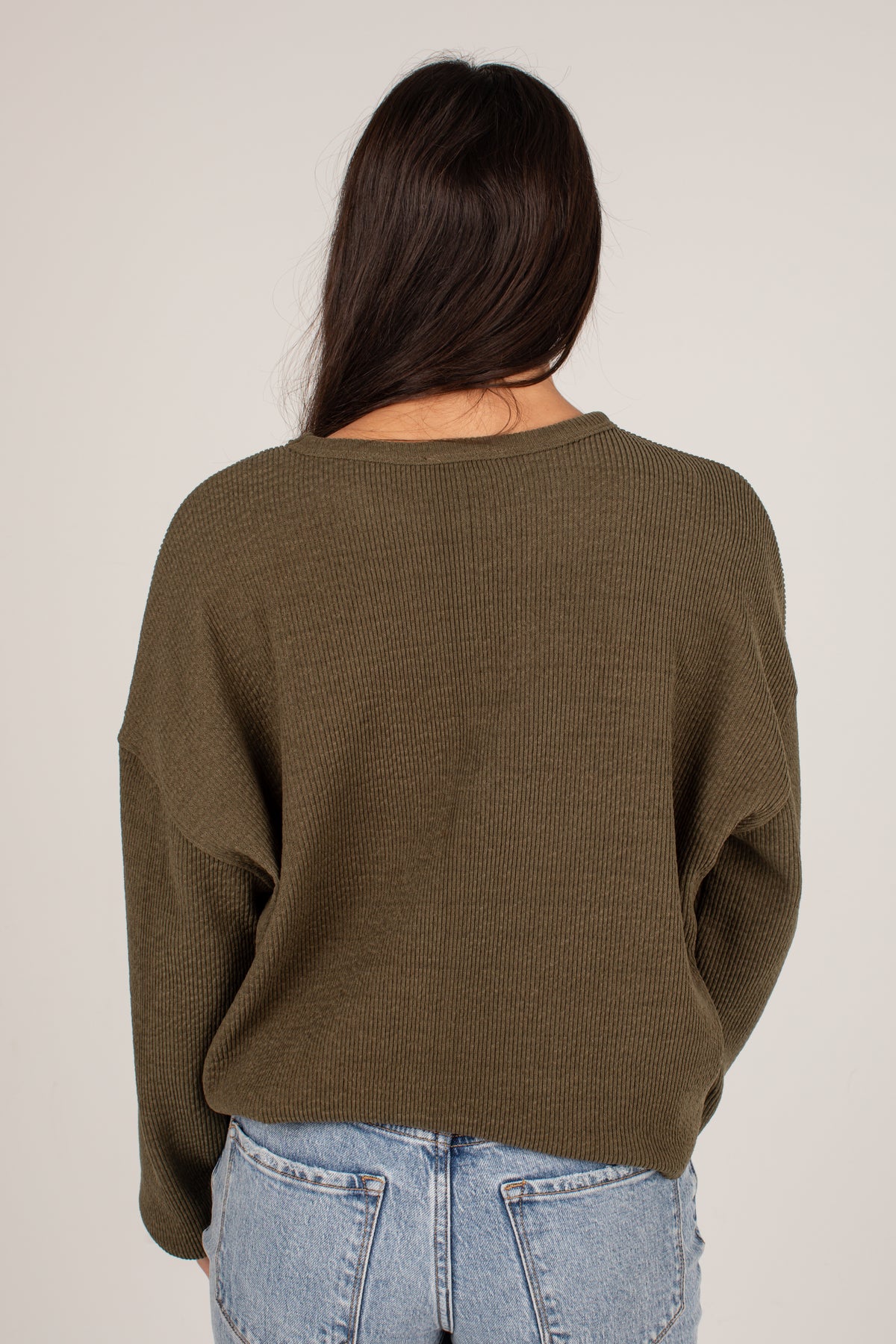 A woman viewed from behind wearing an olive green ribbed knit sweater and light blue jeans against a plain light gray studio background.