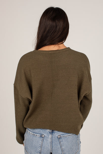 A woman viewed from behind wearing an olive green ribbed knit sweater and light blue jeans against a plain light gray studio background.
