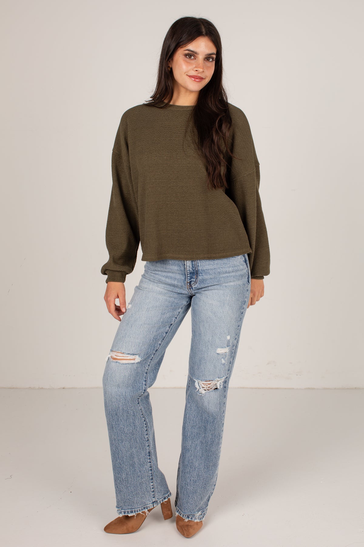 A woman modeling an olive green long-sleeve knit top with light-wash distressed jeans and brown pointed-toe heels, standing against a plain off-white studio background.