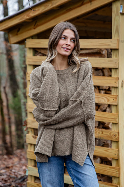 Woman wearing a brown sweater standing in front of a wooden structure with stacked firewood.