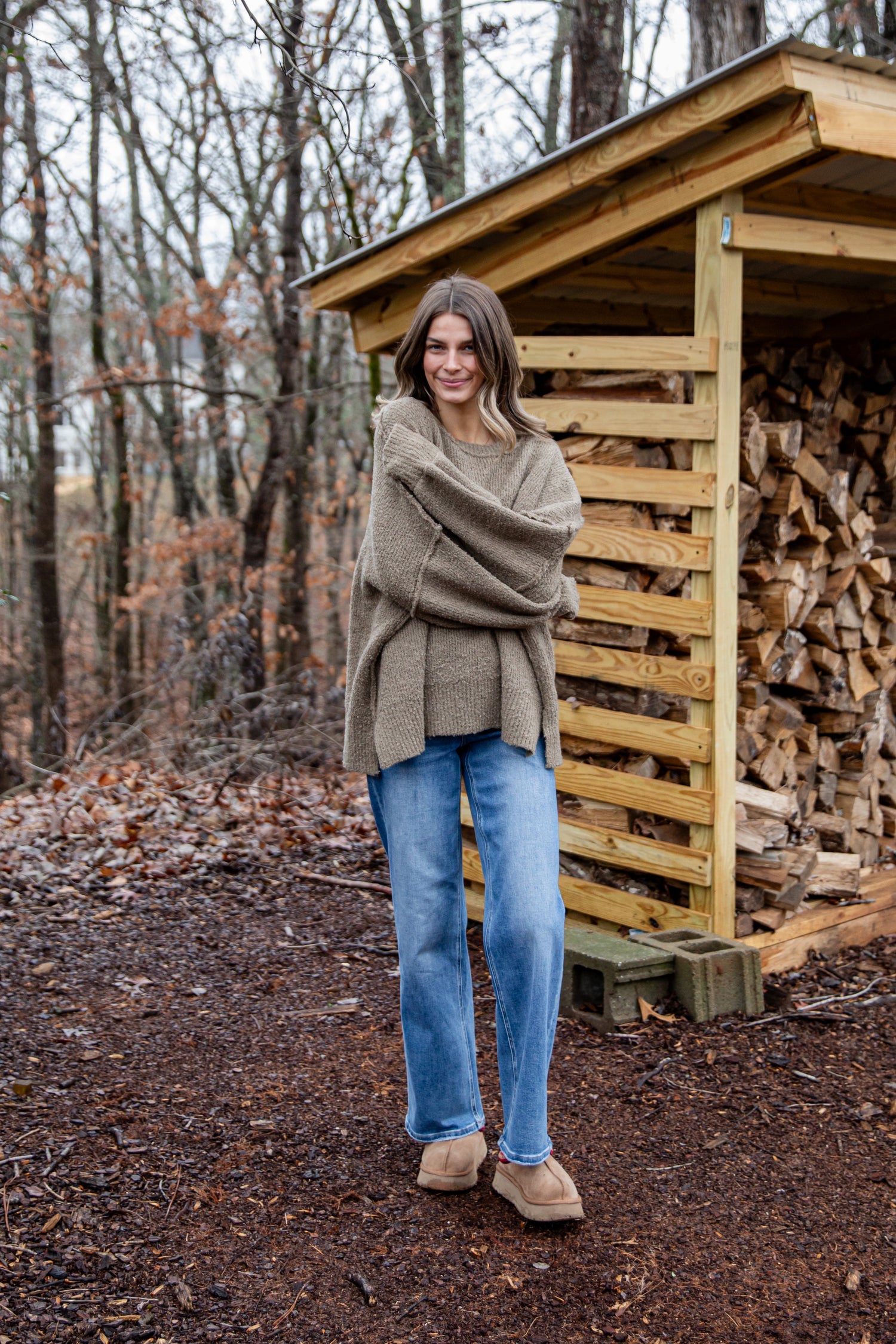 Woman in a beige sweater and blue jeans standing in front of a wooden shed with stacked firewood in a forest setting.