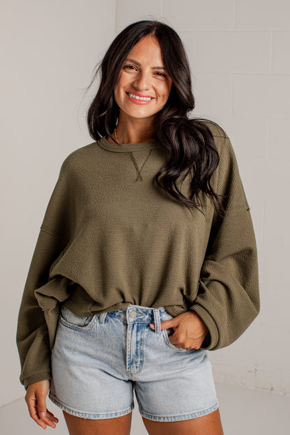 A woman wearing a textured olive green long-sleeve crop sweatshirt and light-wash denim shorts, standing against a light gray brick wall.