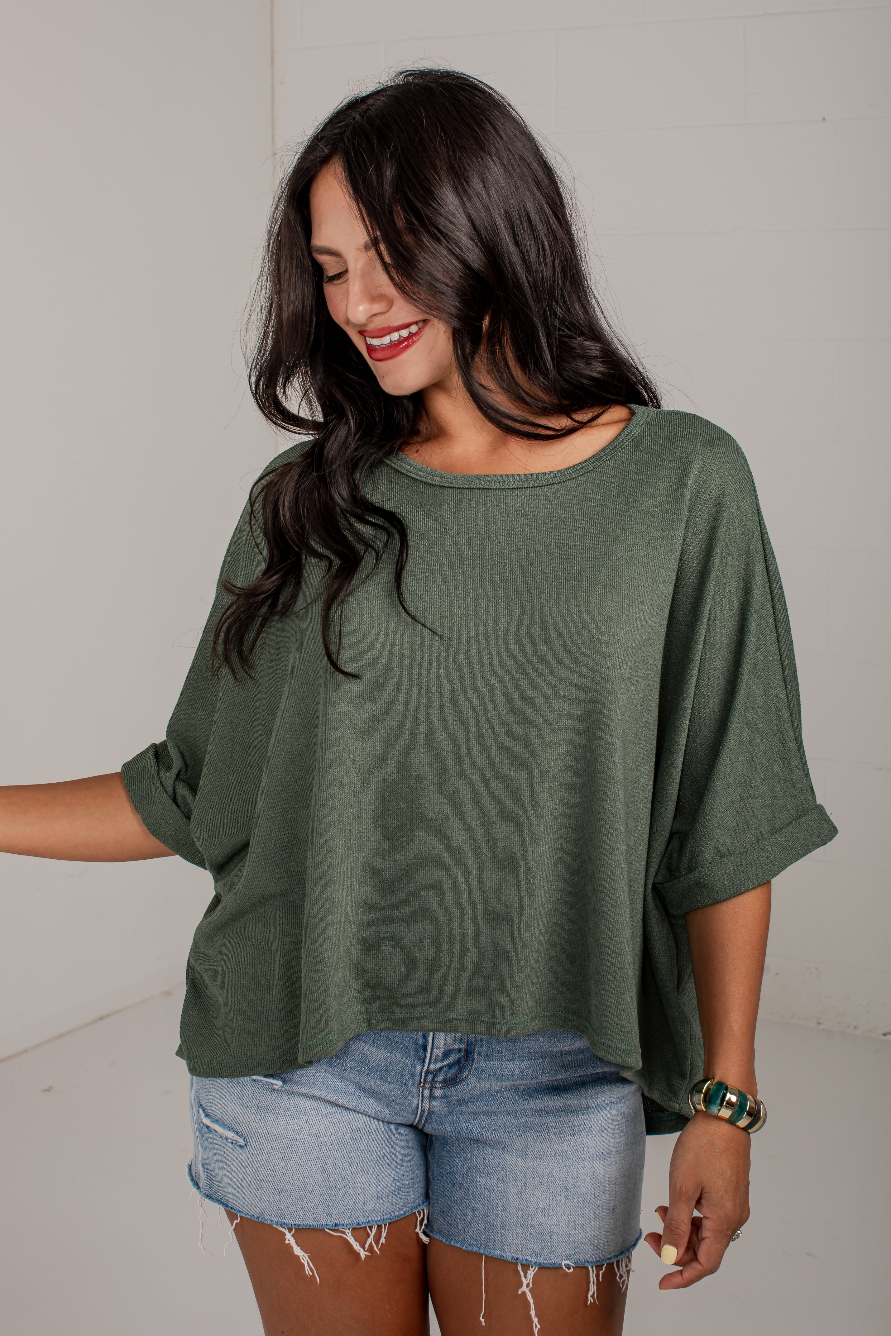 A woman wearing a loose, short-sleeve olive green top and light-wash denim shorts, standing against a plain light gray wall.