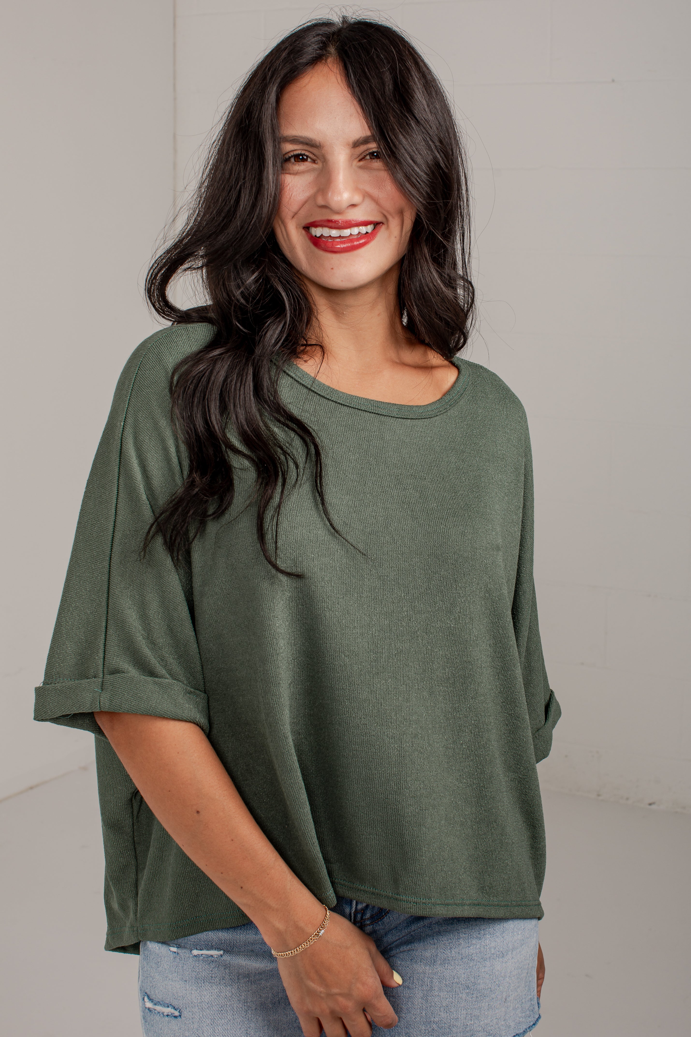 A woman with long dark hair wearing an olive green short-sleeve top and light blue jeans, standing against a plain light gray wall.